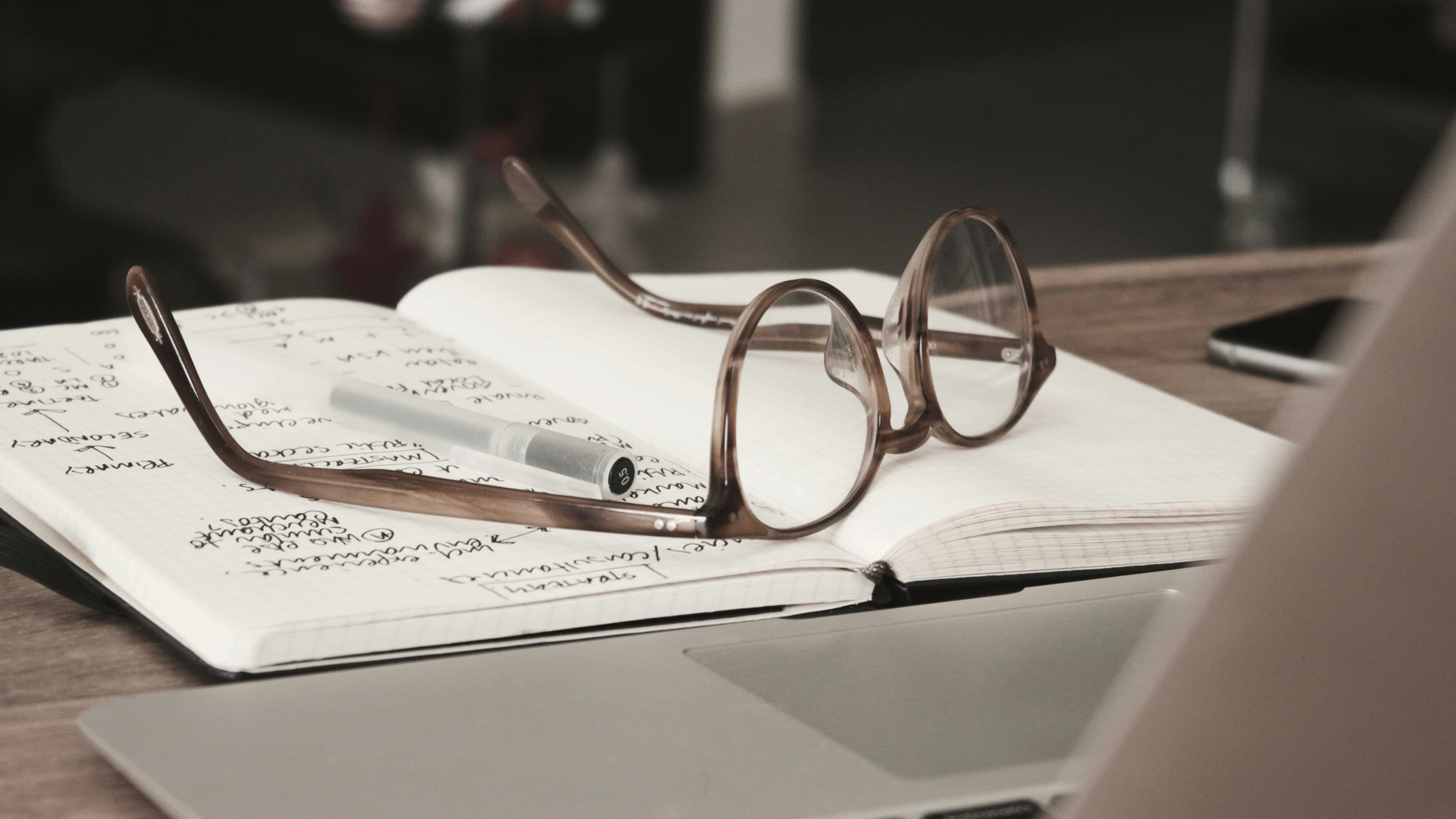 Reading glasses resting on an open notebook of handwritten study notes, beside a laptop.